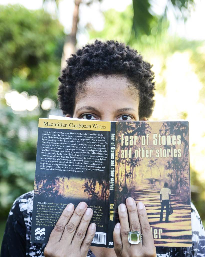 A Black woman (me) is holding Kei Miller's Fear of Stones and Other Stories in front of her face. From her short Afro to her eyes and most of her fingers are visible.